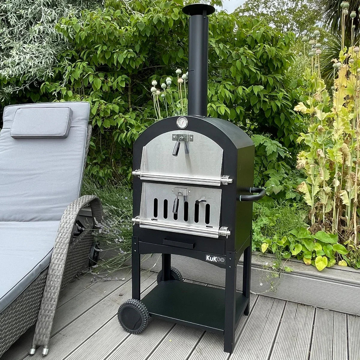 KuKoo outdoor pizza oven with a sleek black finish, stainless steel door, and chimney, set on a wooden patio surrounded by greenery