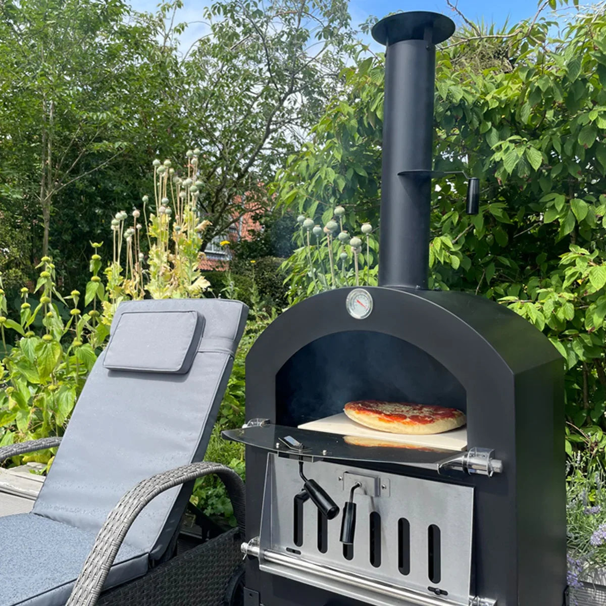 KuKoo outdoor pizza oven with a pizza on the stone, black metal body, chimney, and a garden backdrop with greenery