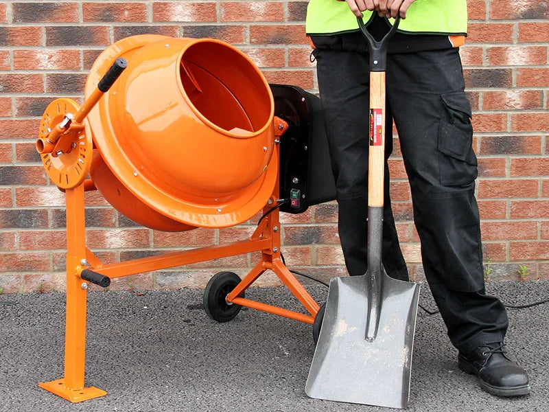 Bright orange concrete mixer with a sturdy metal frame, positioned next to a person holding a metal shovel against a brick wall