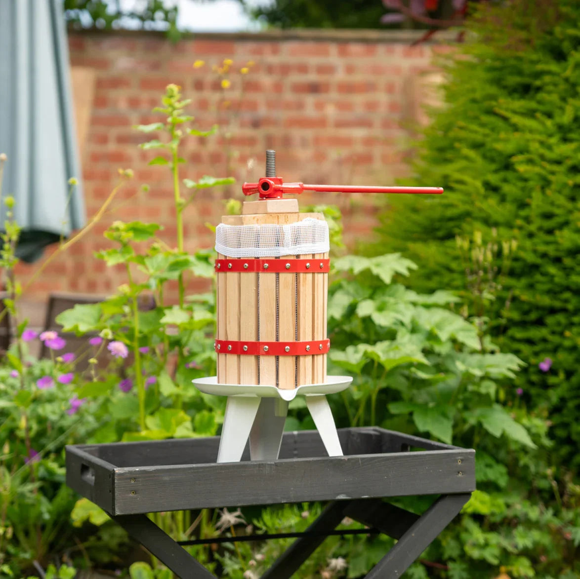 KuKoo 6 Litre Fruit Press with wooden barrel, red metal accents, and a sturdy base, set against a lush garden backdrop