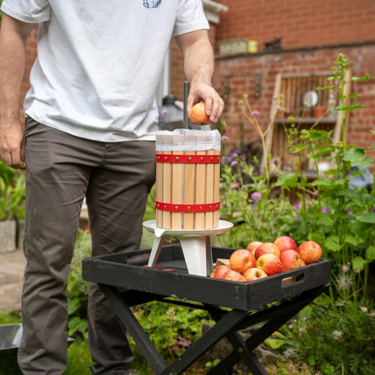 KuKoo 6 Litre Fruit Press with wooden barrel, red straps, and a person adding apples, set in a garden with fresh fruit nearby