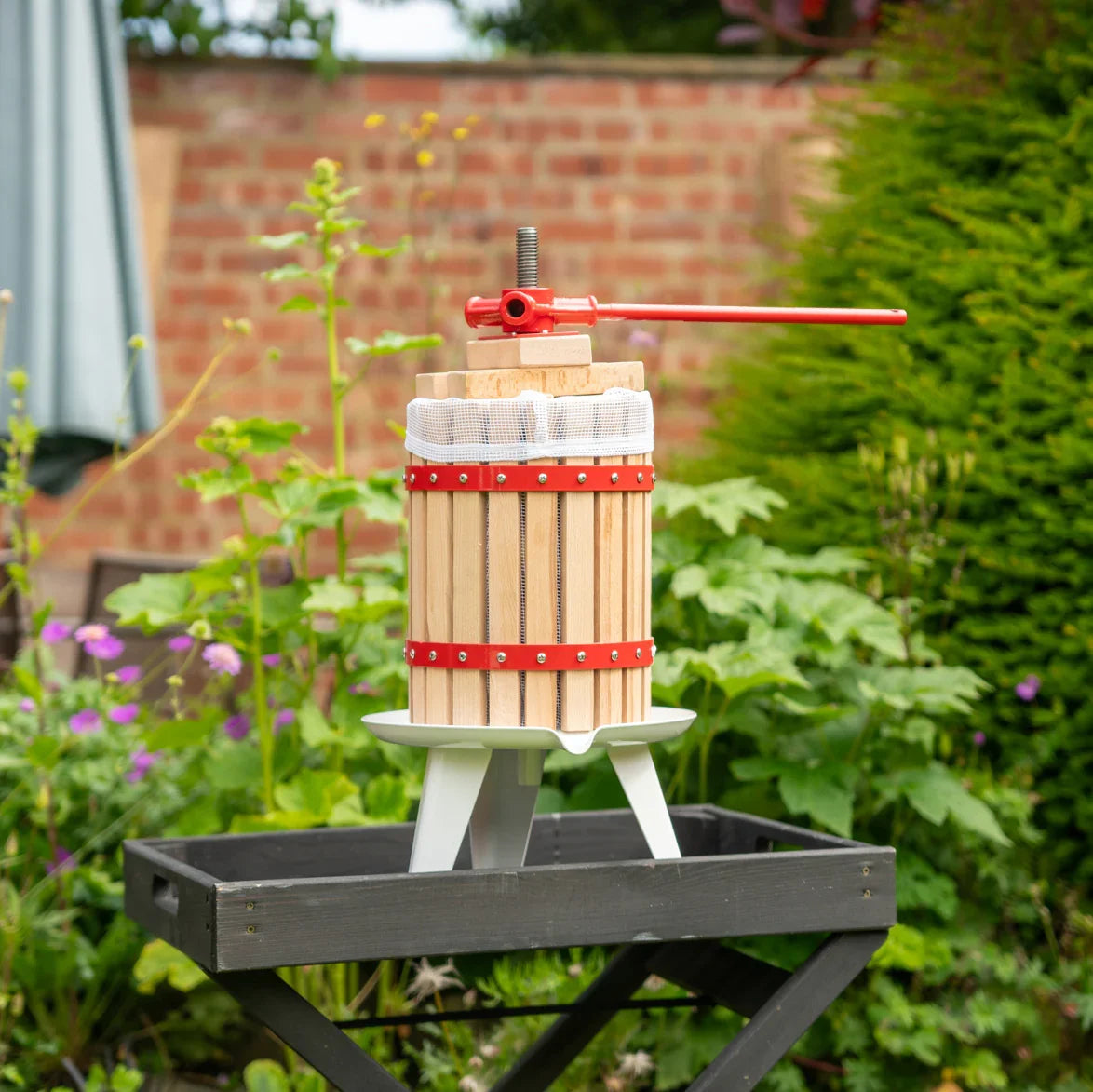 KuKoo 18 Litre Fruit Press with wooden slats, red metal accents, and a sturdy base, set in a vibrant garden with flowers