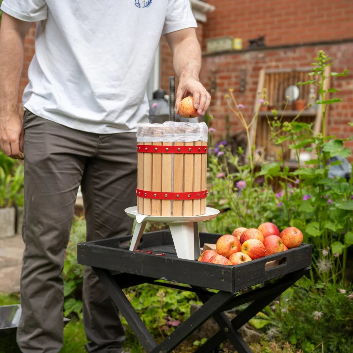 KuKoo 12 Litre Fruit Press with wooden barrel design, red straps, and a person adding apples, set in a garden with fresh apples nearby