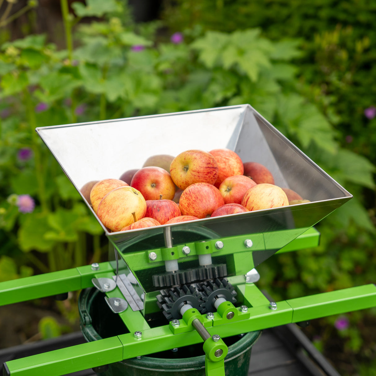Stainless steel fruit crusher with a large hopper filled with red apples, mounted on a green frame for easy fruit processing