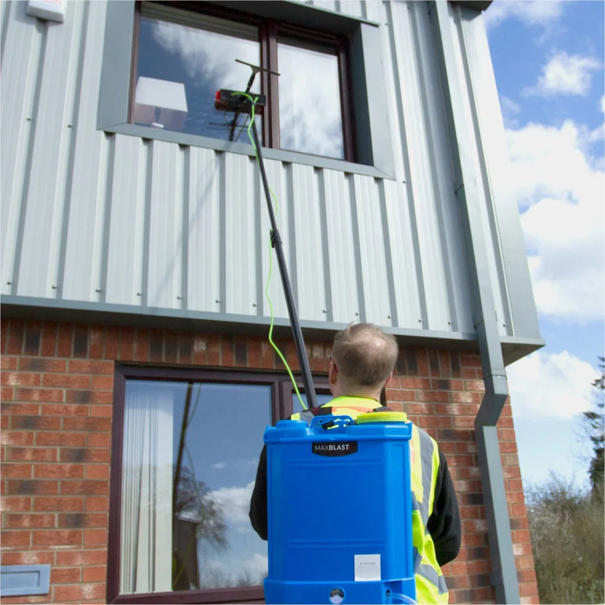 Water fed backpack in blue with 24ft cleaning pole, user cleaning a window on a modern building with cloudy sky background