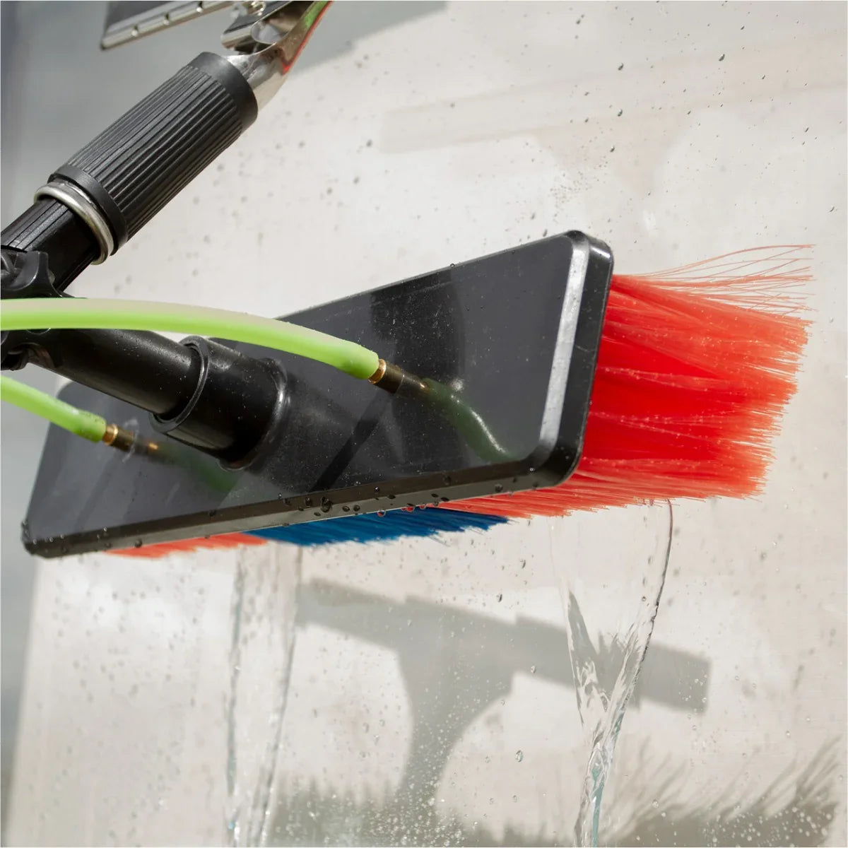 Red and blue bristle brush with green hoses, used for cleaning, attached to a water-fed pole, with water flowing from the...