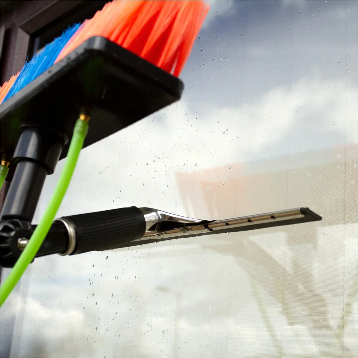 Colorful brush head with soft bristles and squeegee, attached to a water-fed pole, cleaning a glass surface outdoors