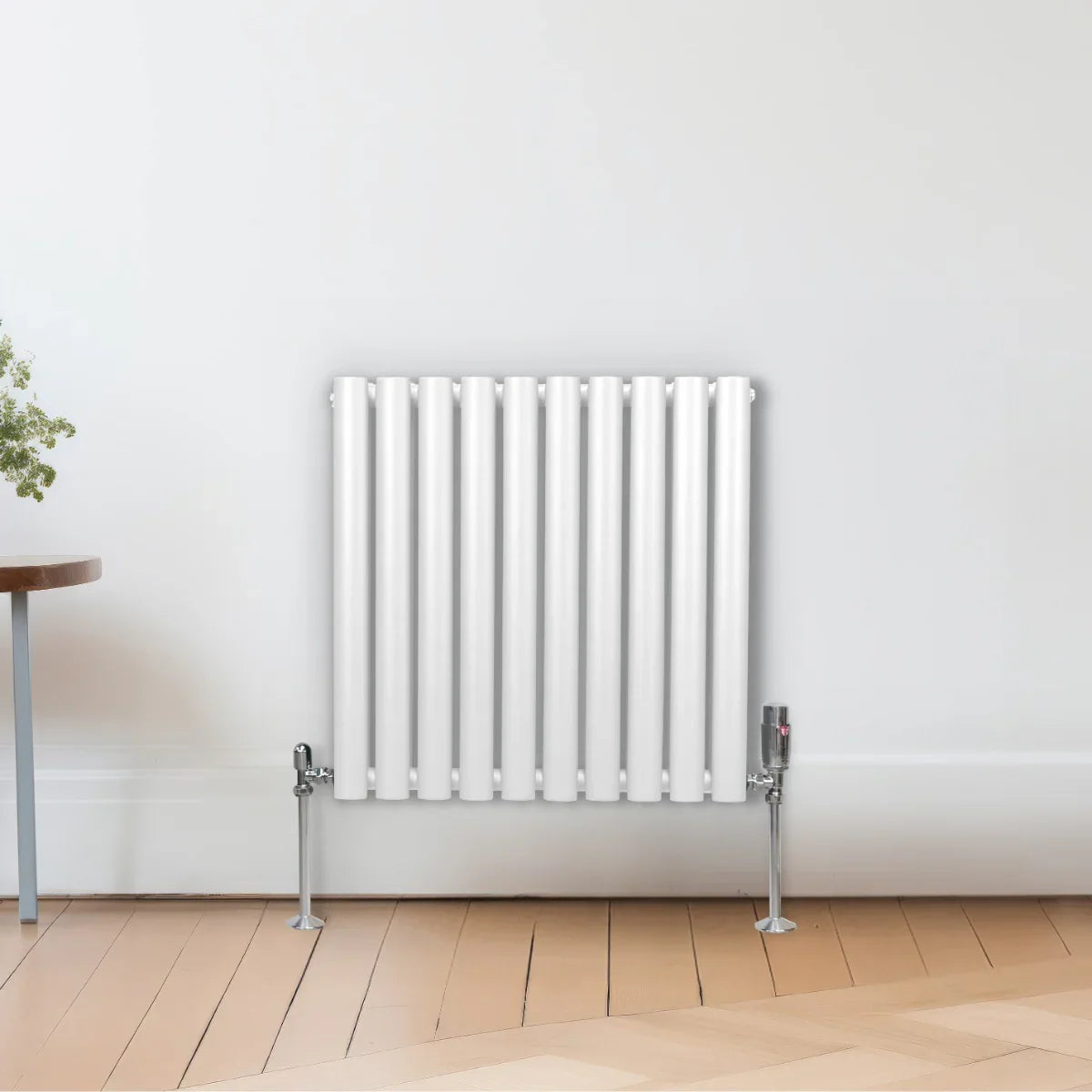 White oval column radiator with vertical fins and chrome valves, mounted on a light wooden floor against a neutral wall