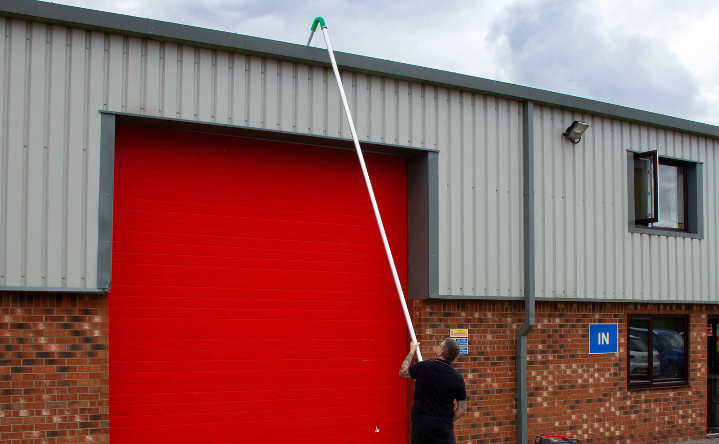 A person using a long pole to reach the top of a red industrial garage door against a gray metal building exterior