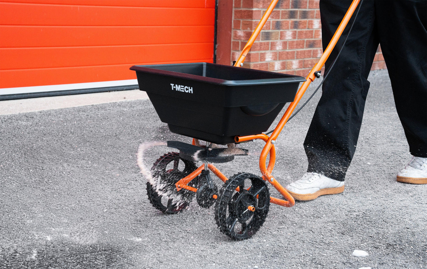 Black T-MECH spreader with orange accents, dispensing white granules on a gray pavement, set against a vibrant red garage door