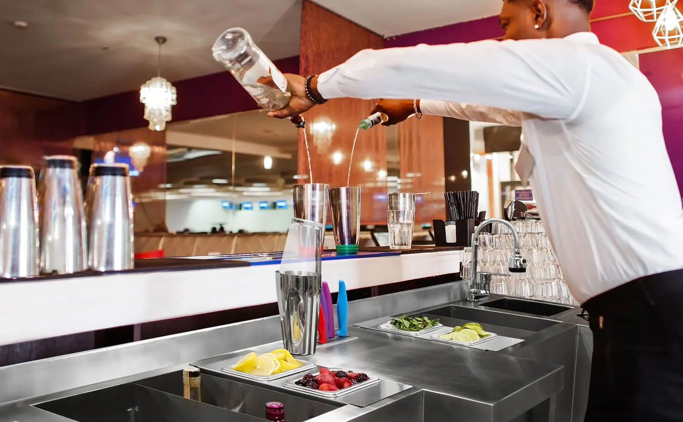 Bartender pouring a cocktail into a shaker, surrounded by fresh fruits, colorful garnishes, and sleek stainless steel bar equipment