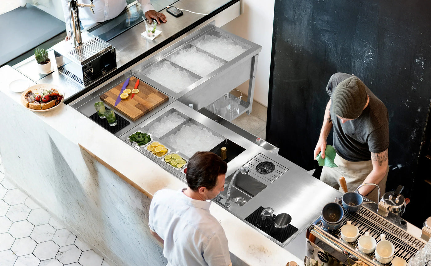 Modern café bar with stainless steel counters, ice bins, fresh ingredients, and a barista preparing drinks in a stylish setting