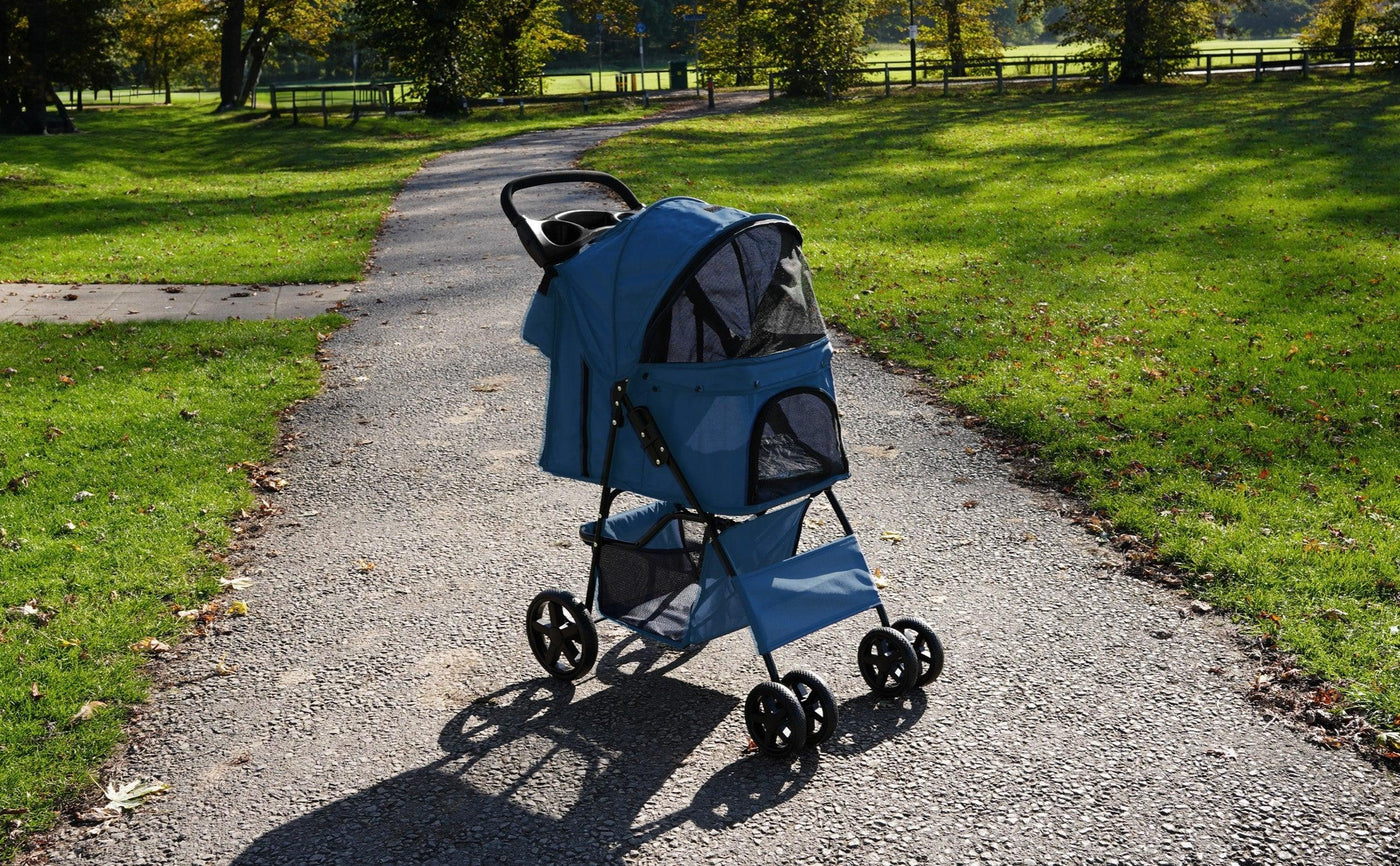 Blue pet stroller with mesh panels and a sturdy frame, parked on a gravel path surrounded by green grass and trees
