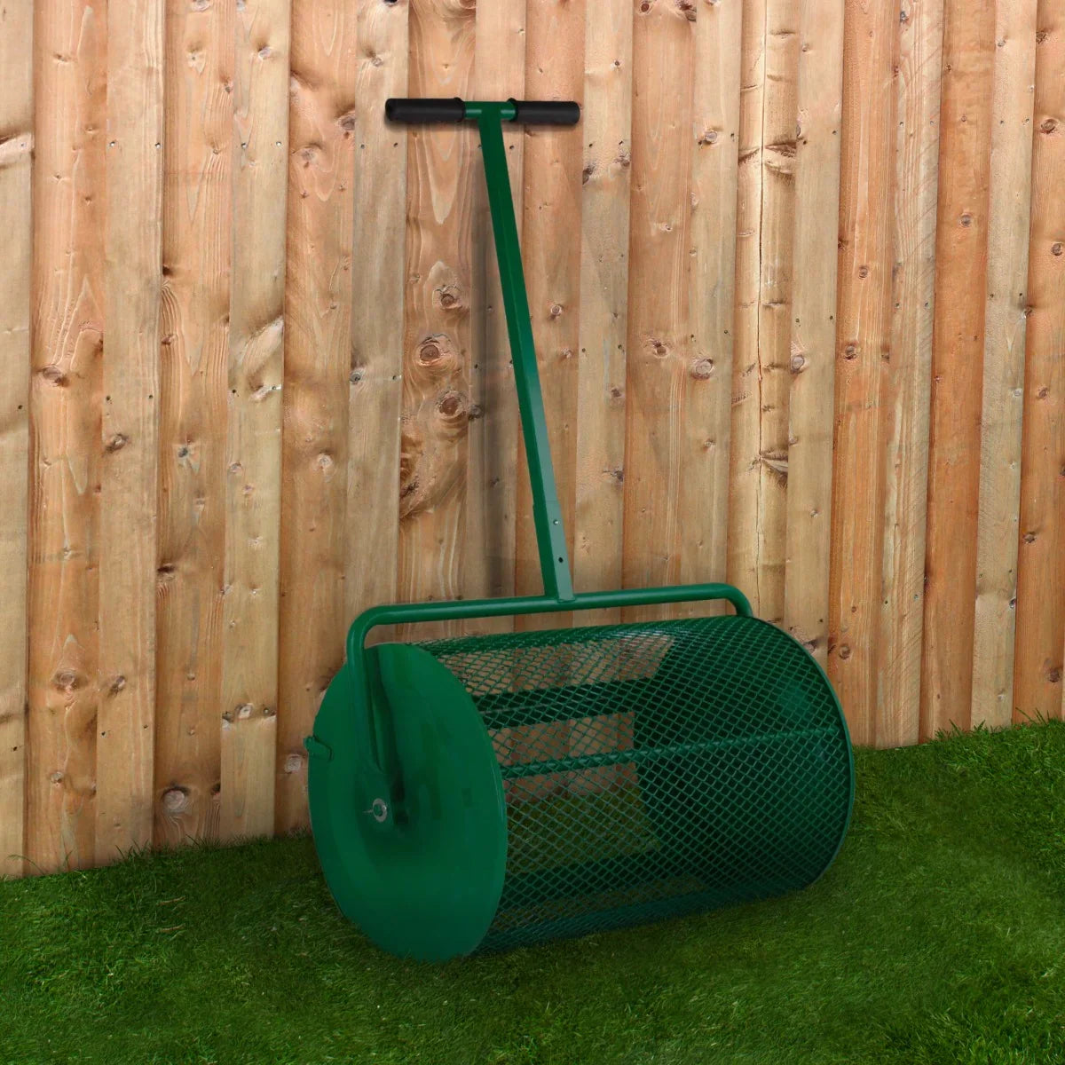Green compost and peat moss spreader with a mesh drum and sturdy handle, positioned against a wooden fence and grass background