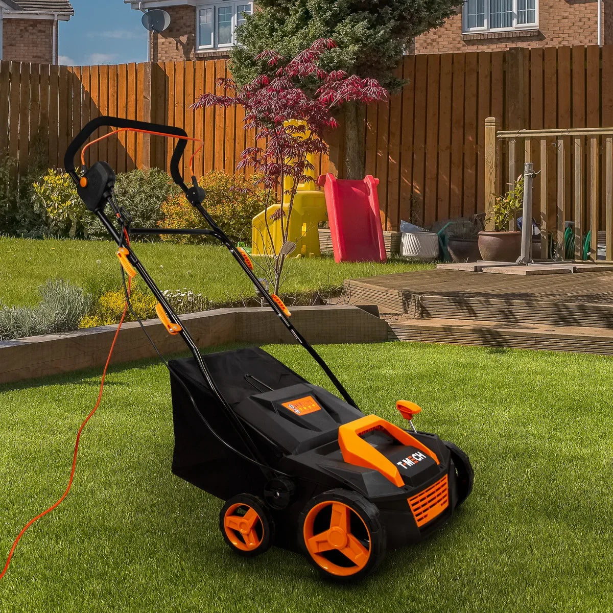 T-Mech Artificial Grass Vacuum with black and orange design, featuring a sturdy handle and wheels, positioned on lush green grass in a garden setting