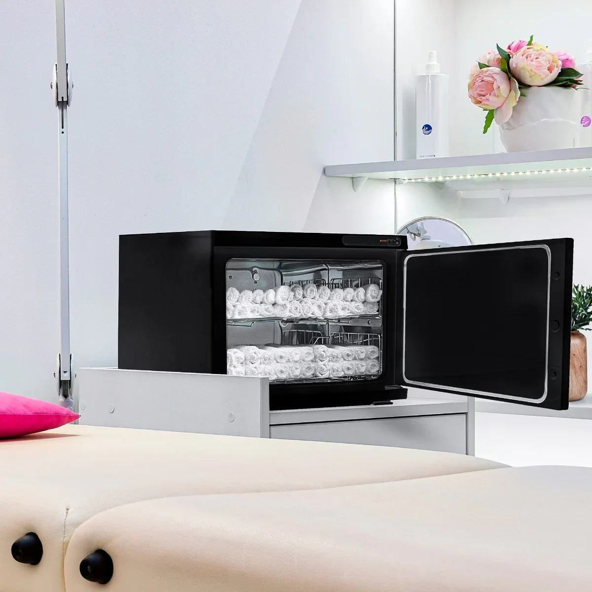Sleek black hot towel warmer with a glass door, filled with rolled towels, placed beside a massage table in a spa setting