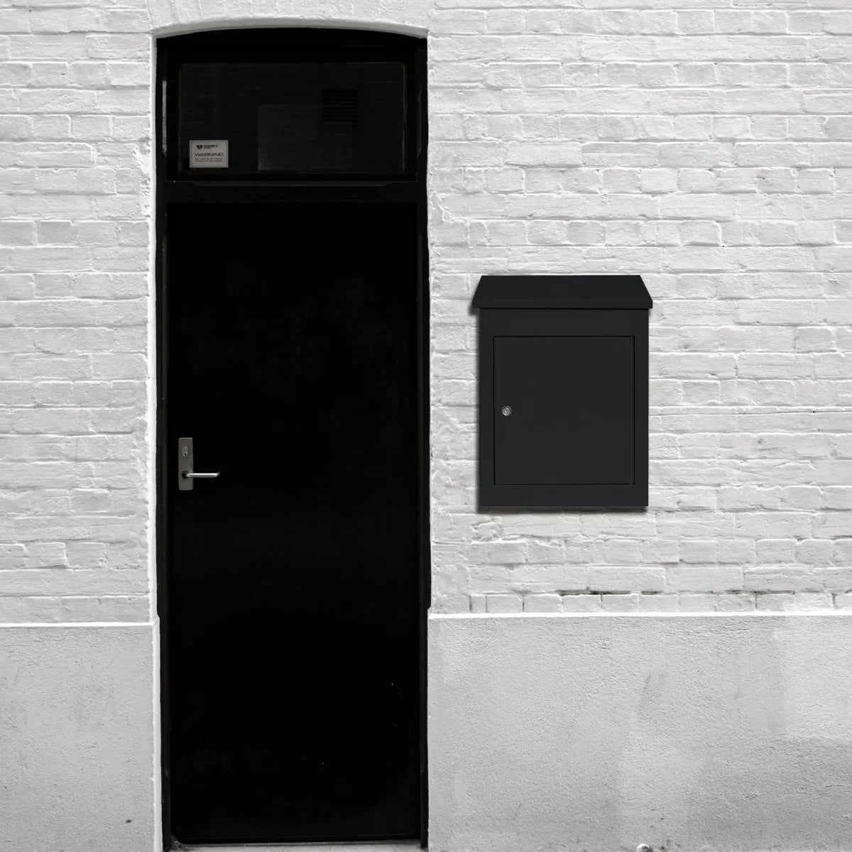 Sleek black anti-theft parcel post box mounted on a white brick wall beside a black door, featuring a secure locking mechanism