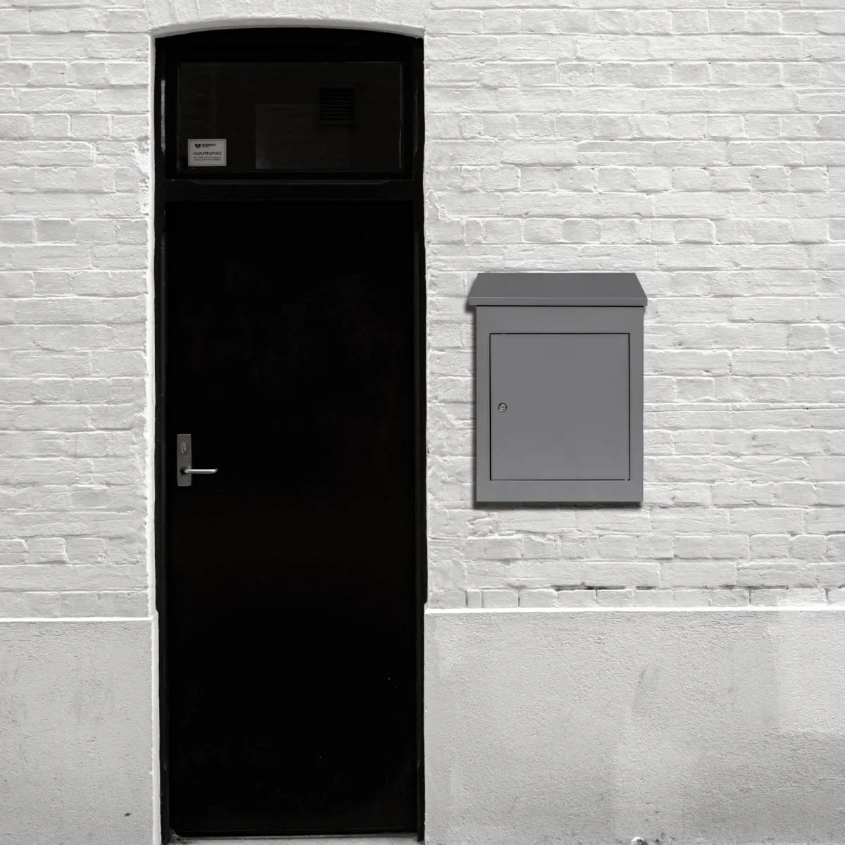 Grey anti-theft parcel post box mounted on a white brick wall beside a black door, featuring a sleek, modern design and secure locking mechanism