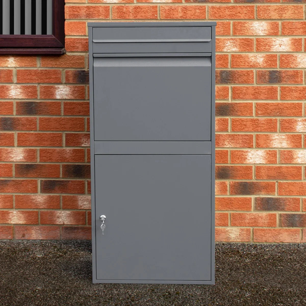 Grey anti-theft parcel post box XL with a sleek design, featuring a top slot and secure lower compartment, set against a brick wall