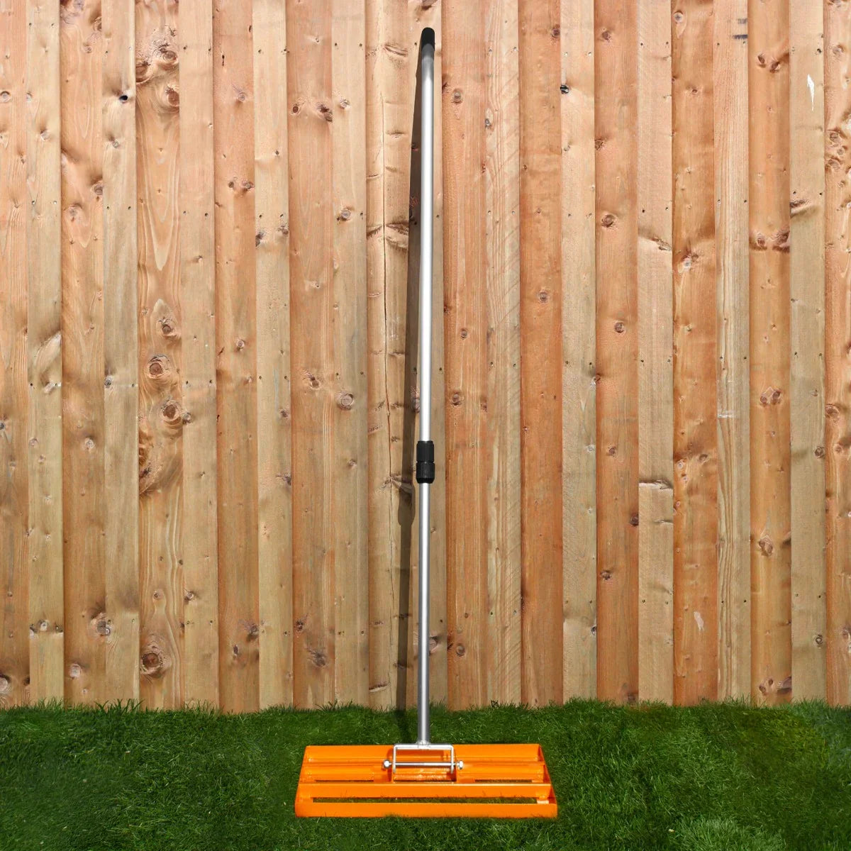 T-Mech Lawn Leveller 50cm in bright orange with a sturdy metal handle, set against a wooden fence and grassy background