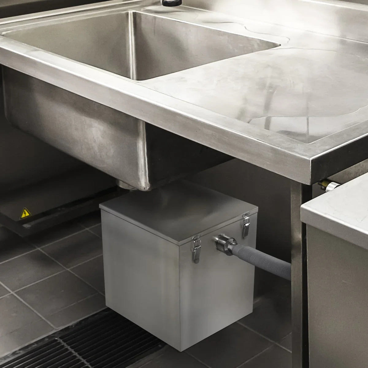 Stainless steel KuKoo grease trap with a 35-litre capacity, installed under a commercial kitchen sink, featuring a drainage hose