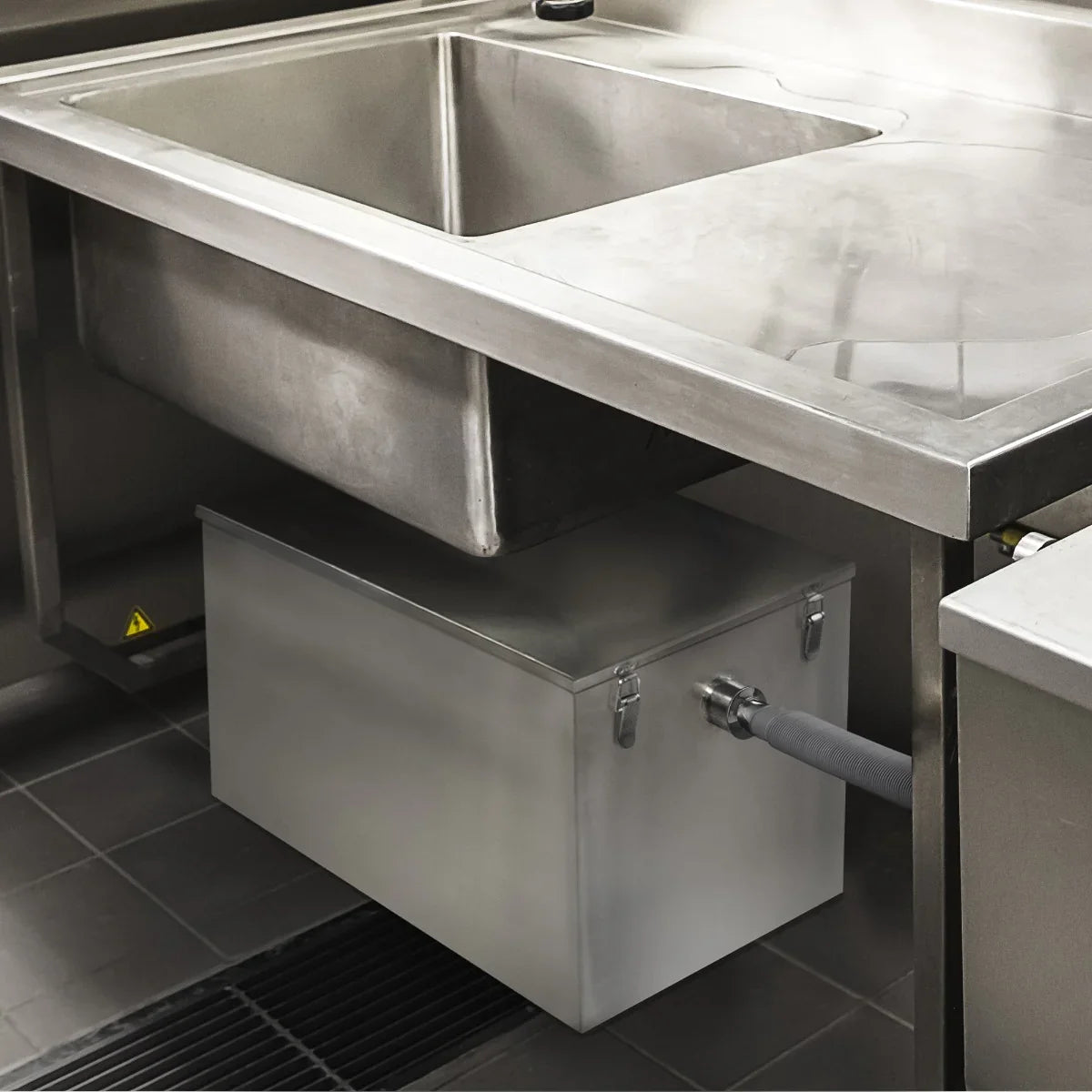 Stainless steel KuKoo grease trap, 120 litres, installed under a commercial kitchen sink with a grey drainage pipe attached