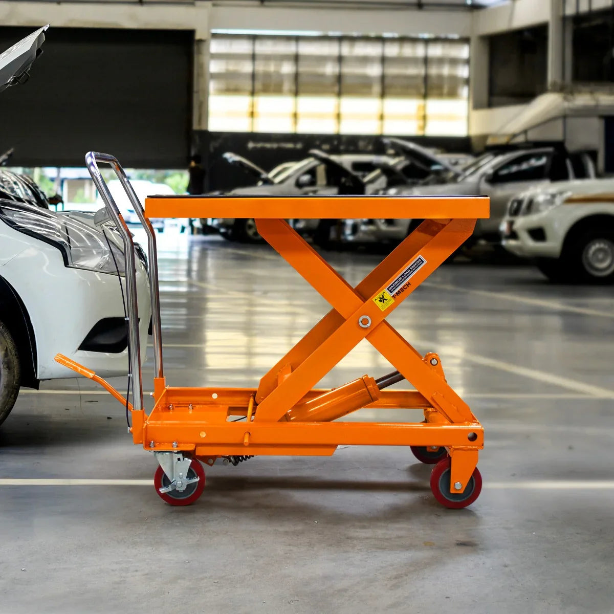 Hydraulic table lift in bright orange, featuring a sturdy steel frame, adjustable height, and smooth-rolling wheels, set in a garage environment