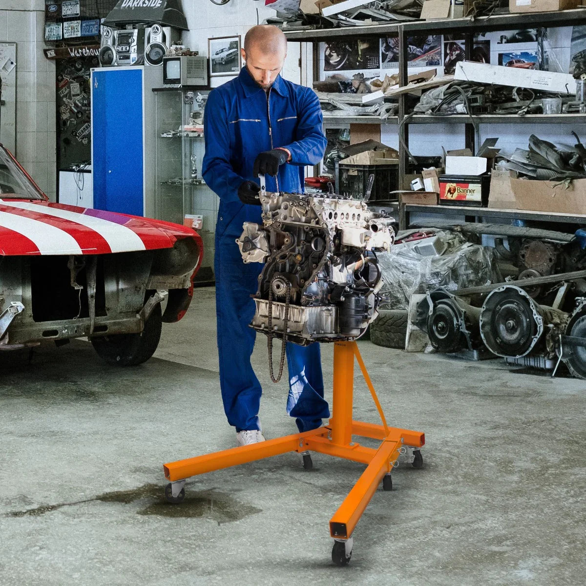 Folding engine stand in bright orange, supporting an engine, with a mechanic in blue coveralls working in a cluttered garage