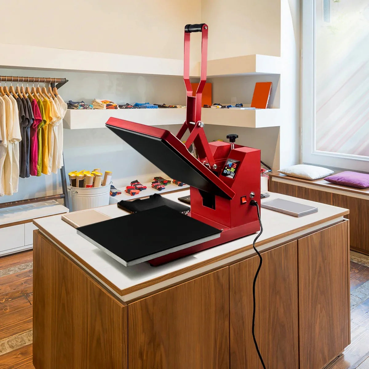 Red Clam heat press with a large black platen, positioned on a wooden table in a bright room with colorful vinyl and shirts in the background