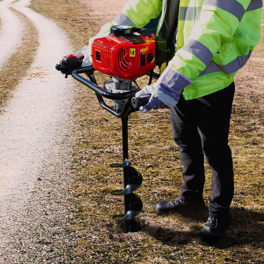A person in a bright yellow safety jacket operates a red gas-powered auger on a gravel path, surrounded by dry grass