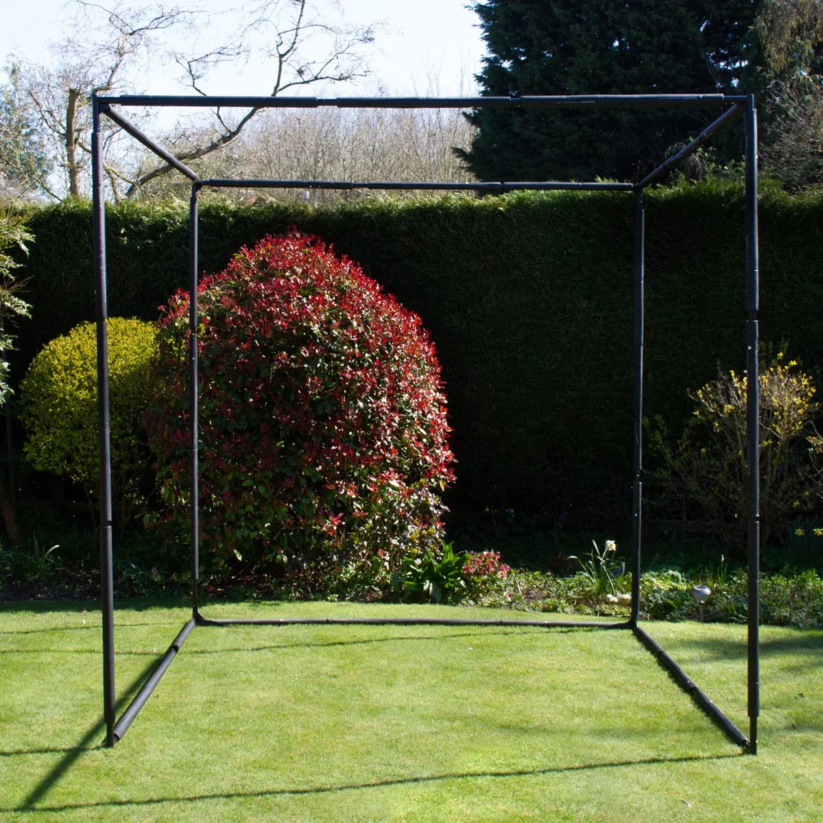 Golf practice cage with a sturdy black frame, set on green grass, surrounded by vibrant shrubs and trees in a sunny garden setting
