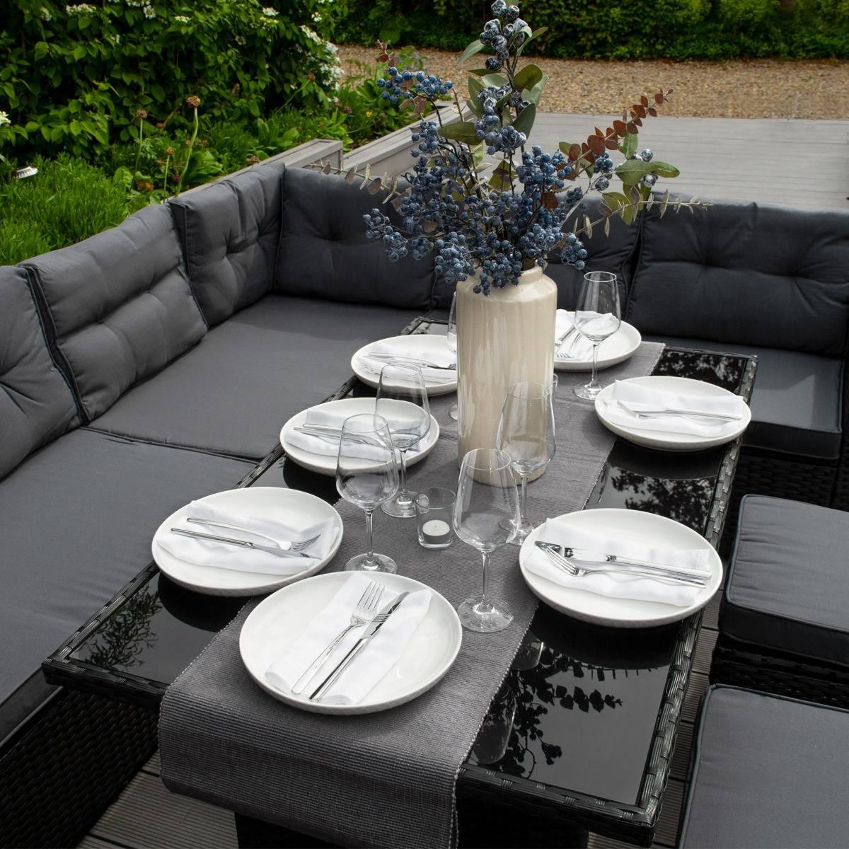 Black rattan corner set with plush gray cushions, elegantly set dining table featuring white plates and glassware, surrounded by greenery