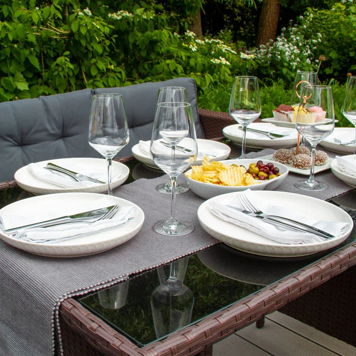 Brown rattan corner set with a glass tabletop, elegantly set with white plates, wine glasses, and a variety of snacks in a lush garden setting