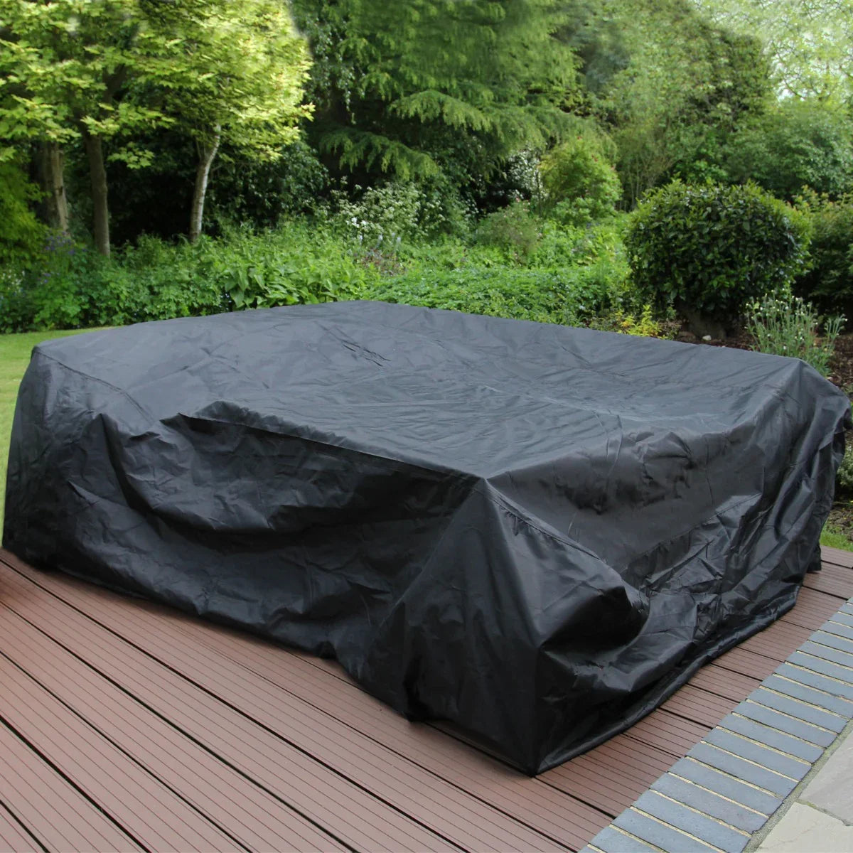 Brown rattan corner set covered with a black protective cover, set on a wooden deck, surrounded by lush greenery and trees