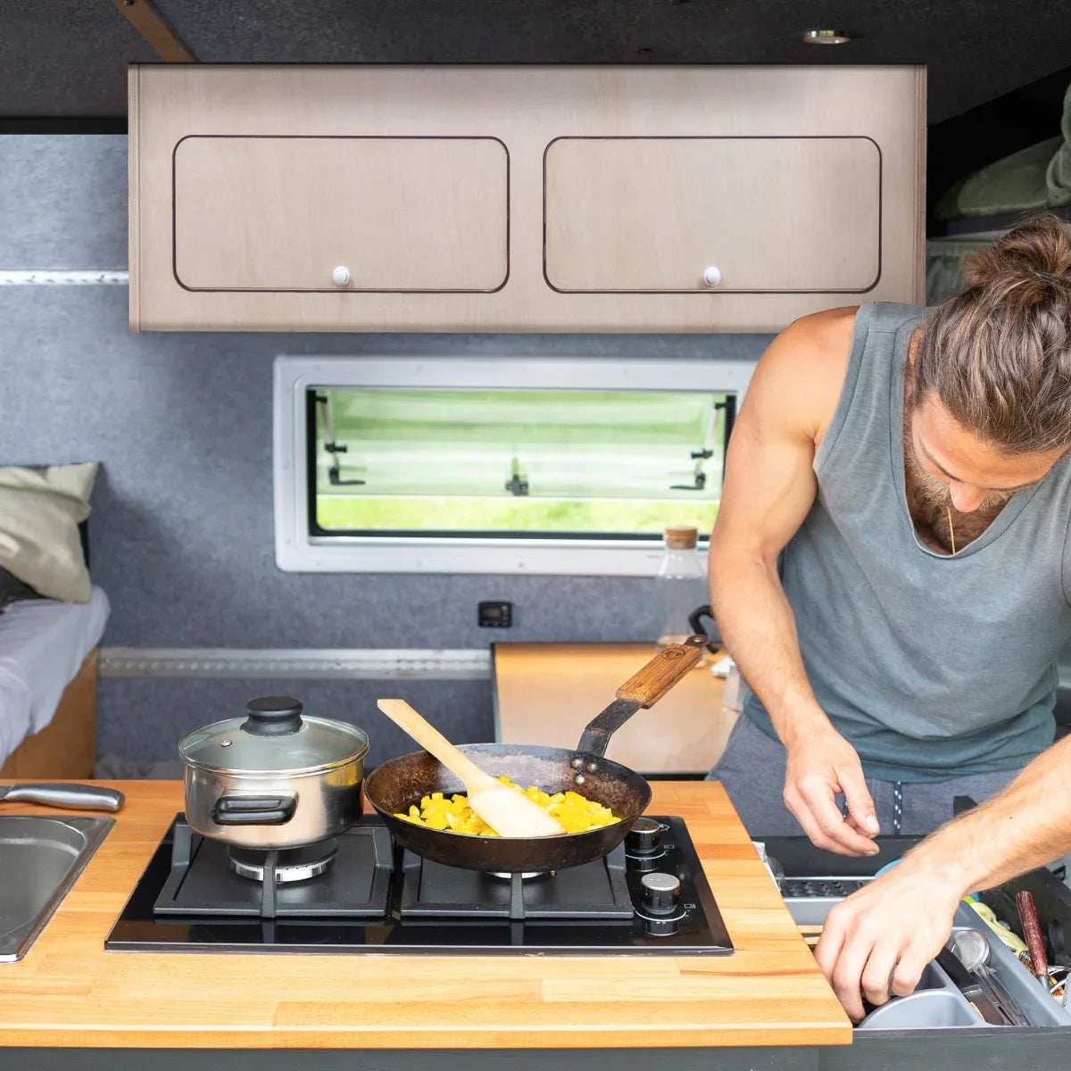 Overhead camper cabinet in light wood finish, featuring two drawers, above a cooking area with a pot and frying pan on a gas stove