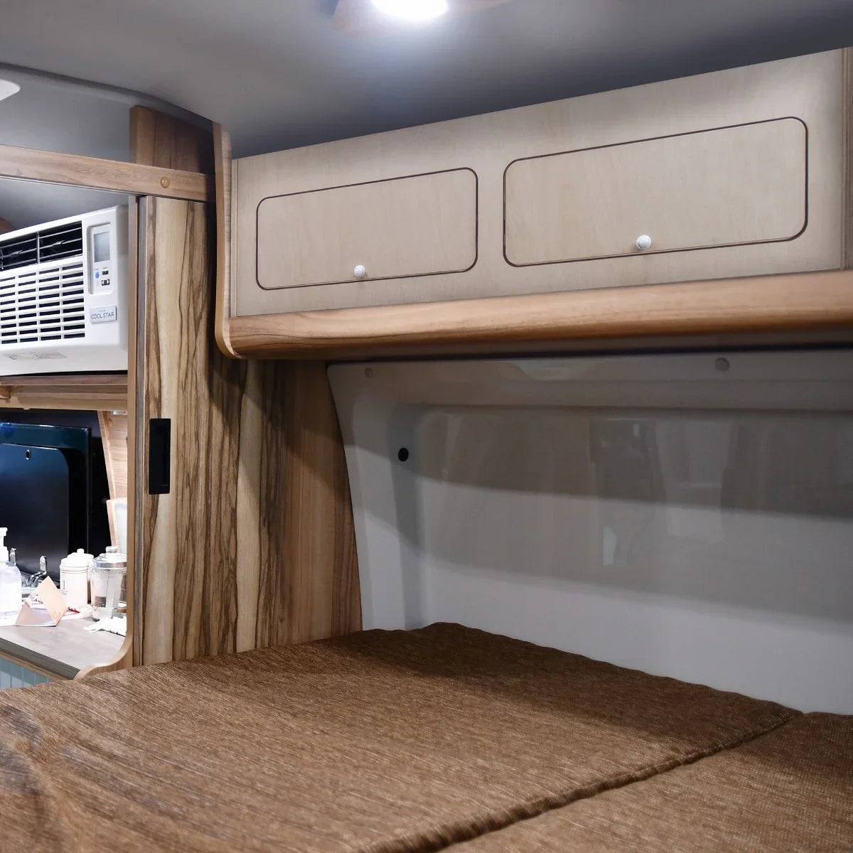Overhead camper cabinet in light wood with two drawers, mounted above a cozy sleeping area, showcasing a sleek, modern design