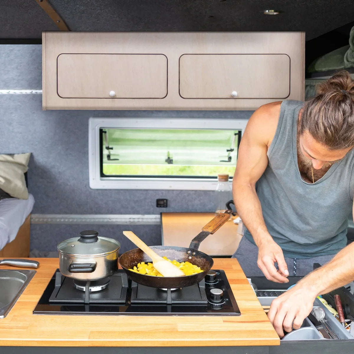 Overhead camper cabinet in light wood, featuring two upper storage compartments, above a compact kitchen with cooking utensils and food preparation