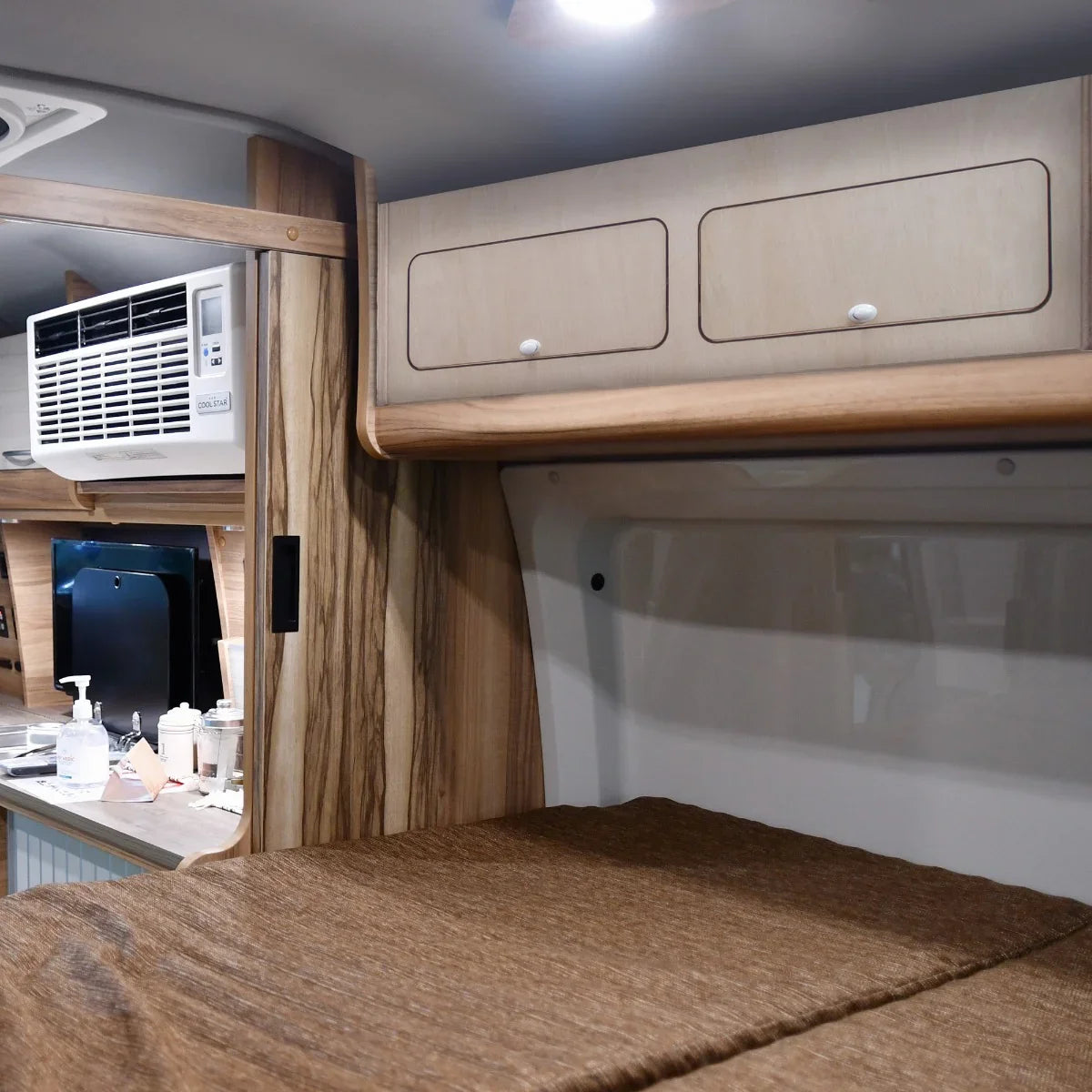Overhead camper cabinet in light wood finish with two drawers, mounted above a cozy sleeping area, featuring a soft brown blanket