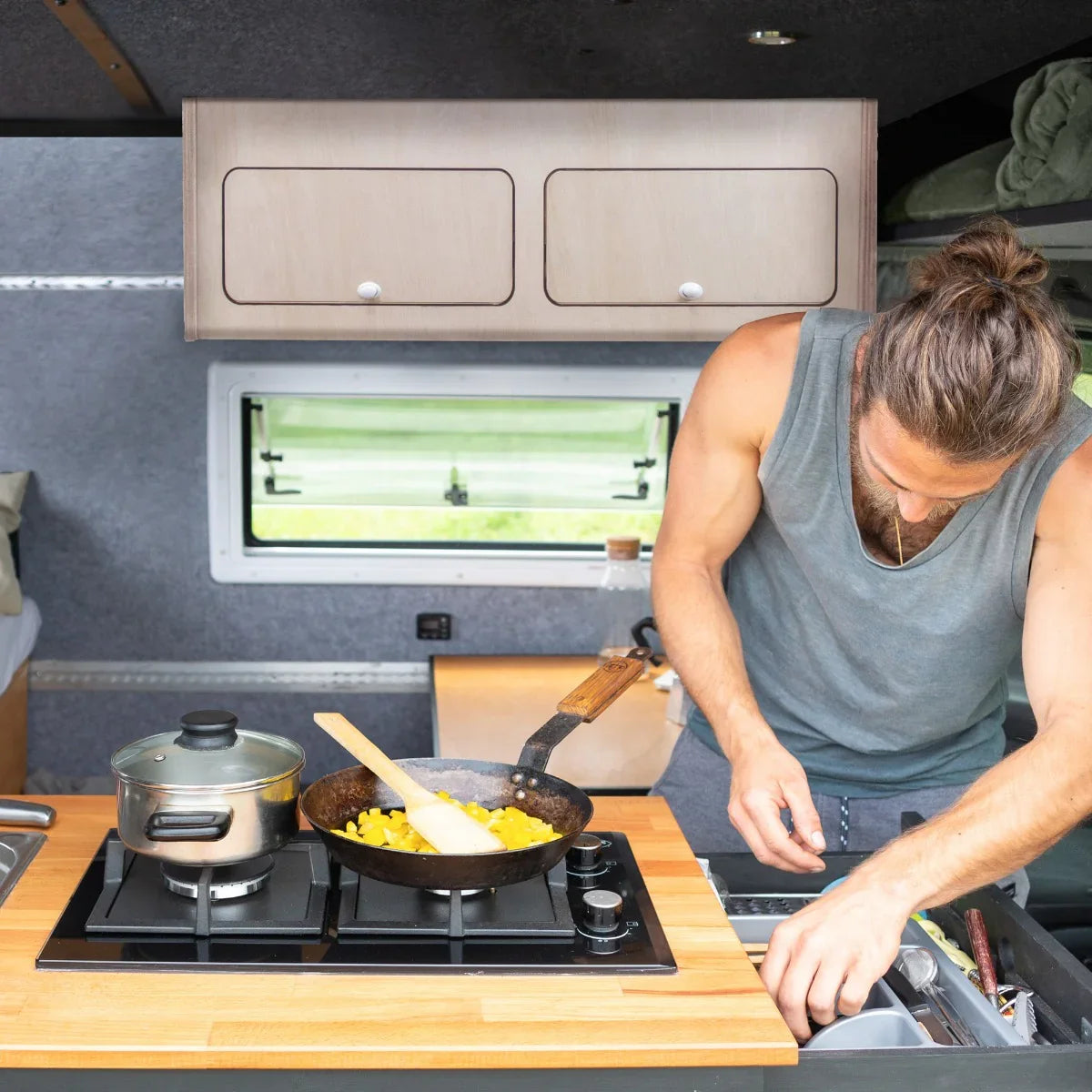 Overhead camper cabinet in light wood finish, featuring two drawers, above a compact kitchen with a man cooking on a stove