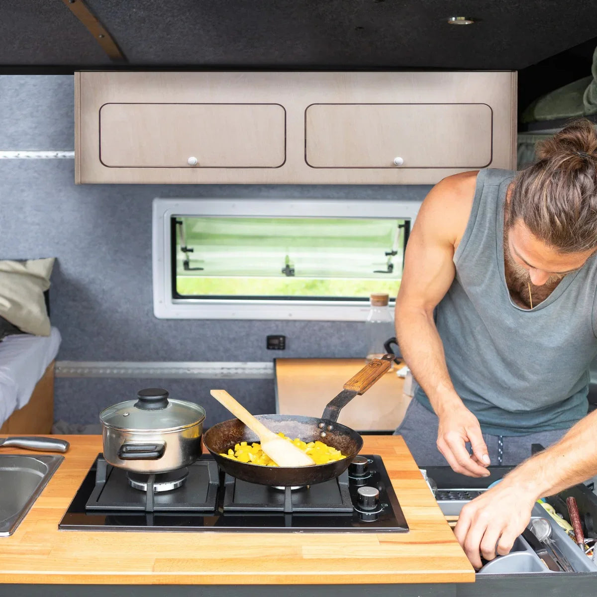 Overhead camper cabinet in light wood, mounted above a kitchen area with a person cooking on a gas stove, featuring a modern design