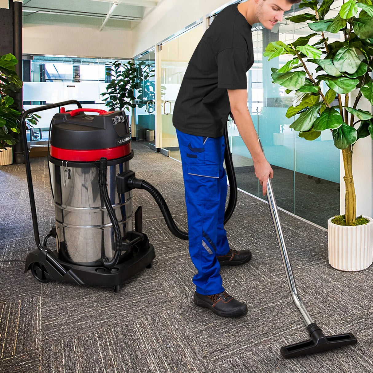 80L industrial vacuum with stainless steel body, red accents, and flexible hose, used by a person in blue workwear in a modern office setting