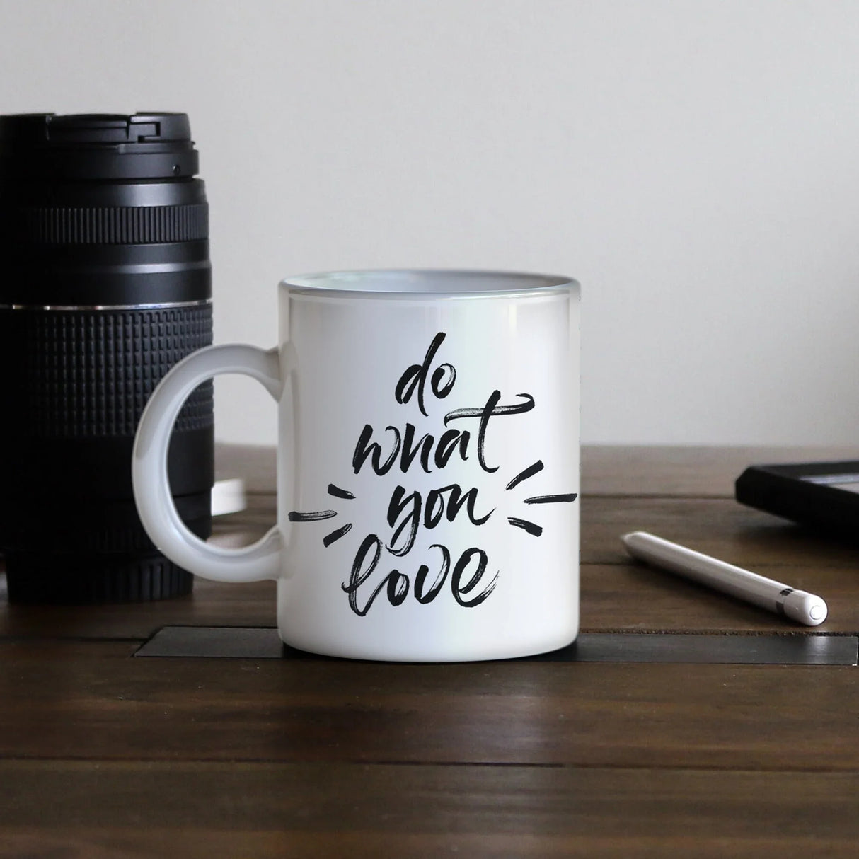 White ceramic mug with black script "do what you love" on wooden surface, camera lens and stylus nearby.