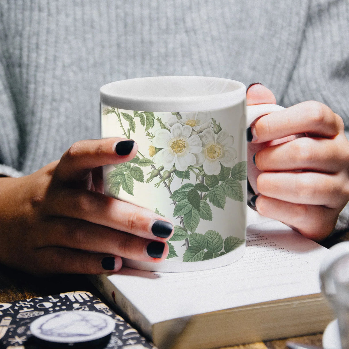 White ceramic mug with floral design held by hands over an open book.