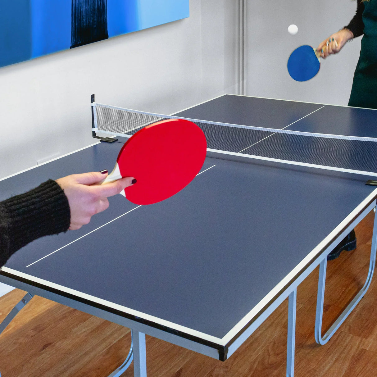 Folding table tennis table with blue surface, net, and red and blue paddles in action, set in a bright indoor space