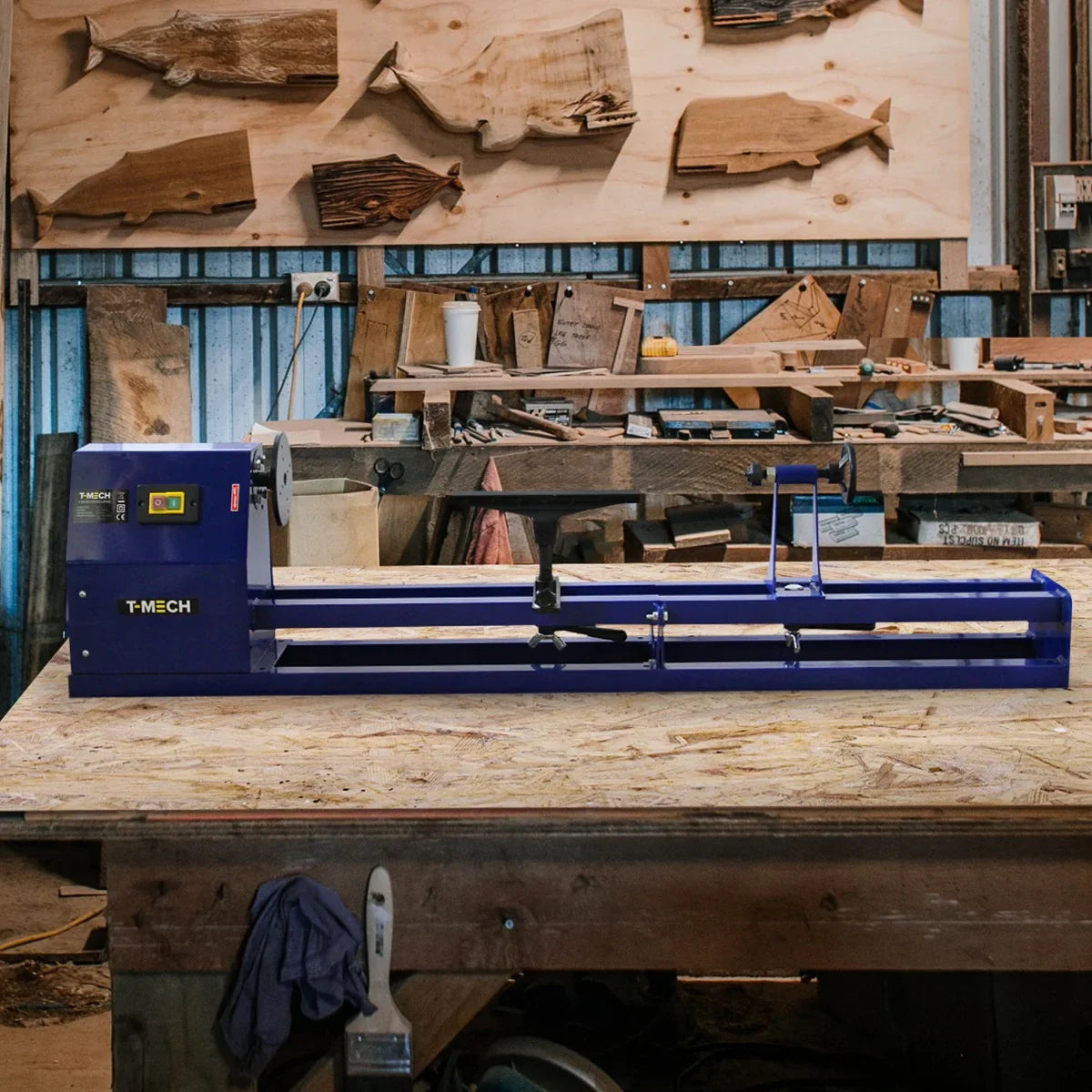 Blue wood lathe with a 4 jaw chuck, set on a wooden workbench surrounded by various wood pieces and tools in a workshop setting