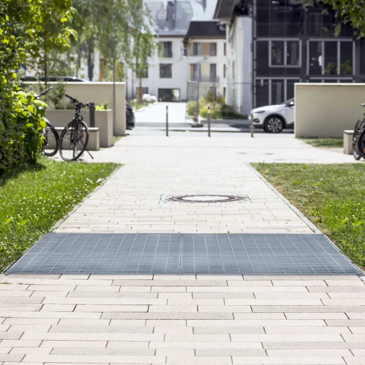 Galvanised grating measuring 1200mm x 1000mm, featuring a sturdy metal grid design, placed on a paved walkway surrounded by greenery