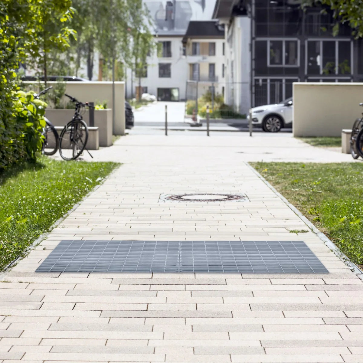 Galvanised grating measuring 500mm x 500mm, featuring a sturdy grid design, set on a paved walkway with greenery and buildings in the background
