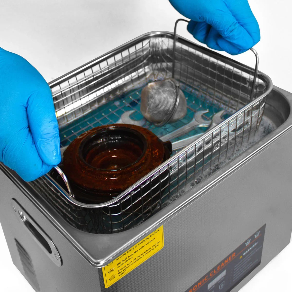 Hands wearing blue gloves lifting a metal basket from the T-Mech Ultrasonic Cleaner, revealing a brown ceramic pot and cleaning tools submerged in water