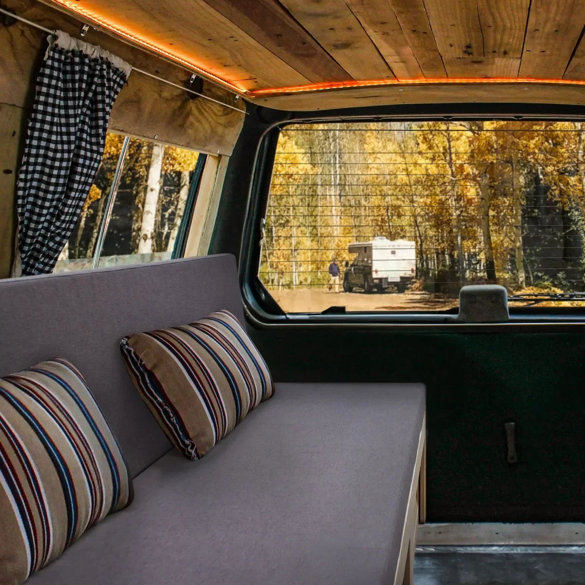 Cozy camper van bed with gray upholstery and striped pillows, featuring wooden accents and a scenic forest view through the window