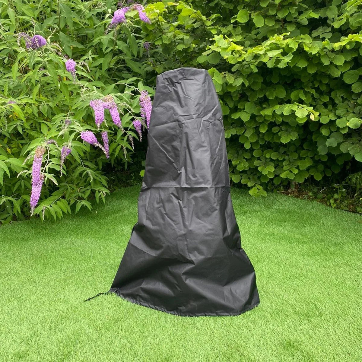 Black protective cover for a cast iron chimenea, designed to shield it from weather elements, set against a lush green garden backdrop